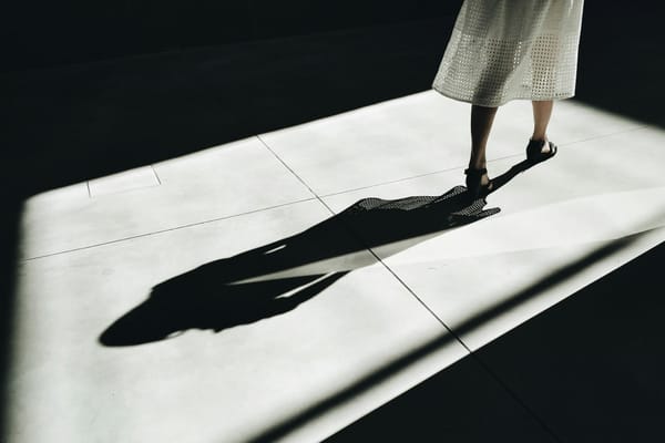 Kaki perempuan bergaun putih melangkah di lantai yang terang, bayangannya jatuh panjang ke area gelap